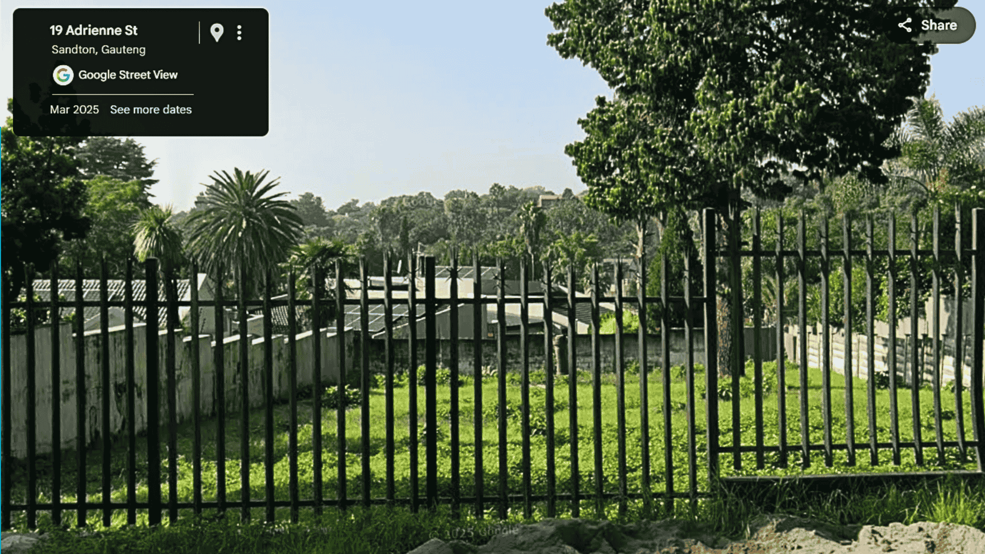 A tall black fence encloses a grassy yard with scattered trees and a few palm trees, reminiscent of the Magaliesberg region. Behind the fence are more trees and low concrete buildings, all bathed in sunlight.