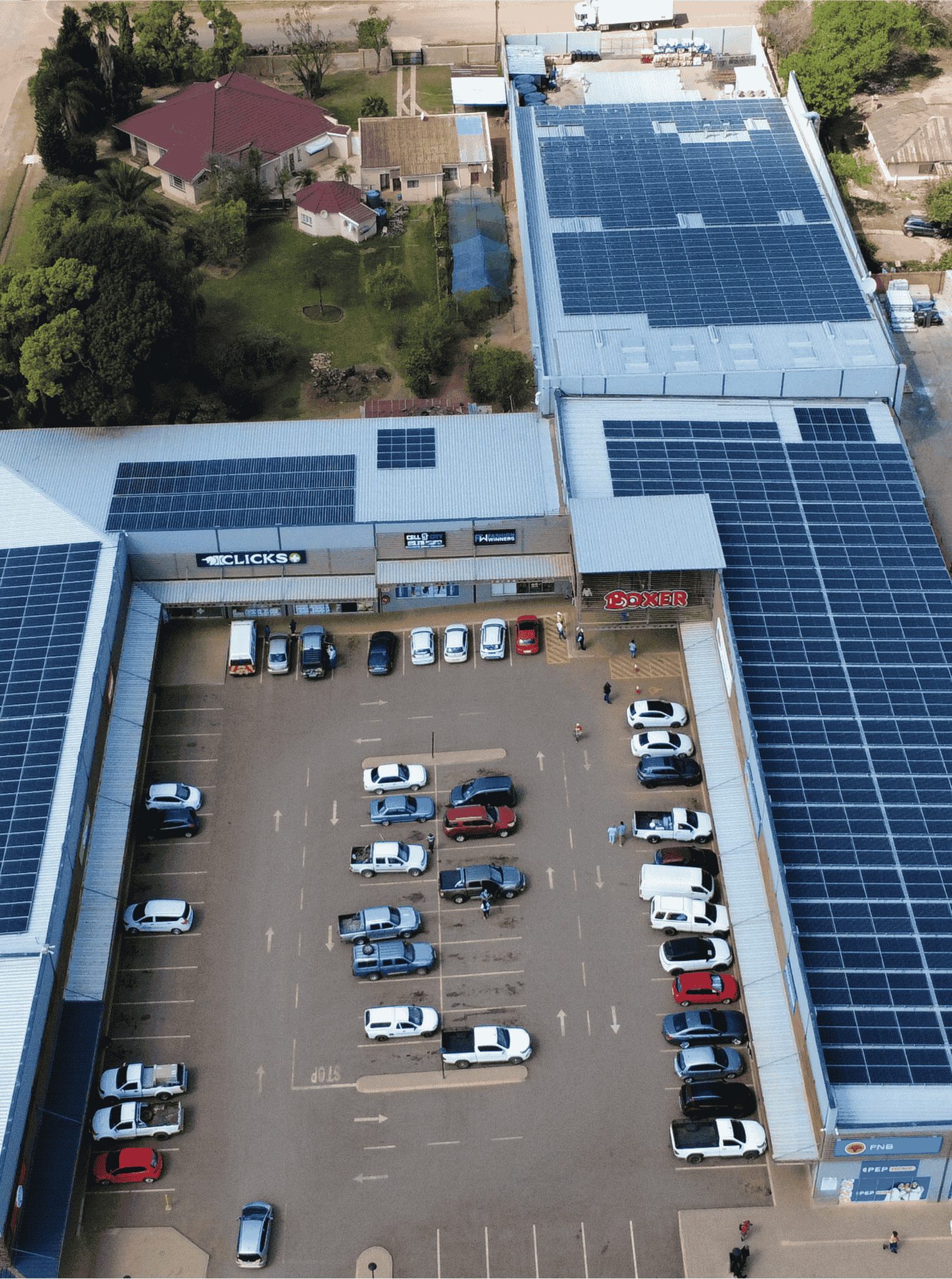 Aerial view of a parking lot outside a shopping center with numerous parked cars. Solar panels cover the building roofs, revealing smart business opportunities for sustainable energy. A few trees and houses appear in the background.