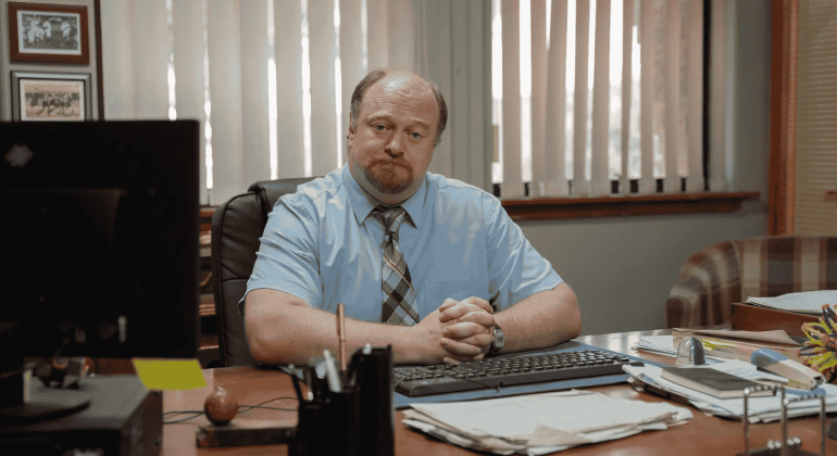 A man with a beard and mustache, wearing a light blue shirt and striped tie, sits at his desk with hands folded. He faces the camera in an office with window blinds, various papers, and notes about road closures on the desk.