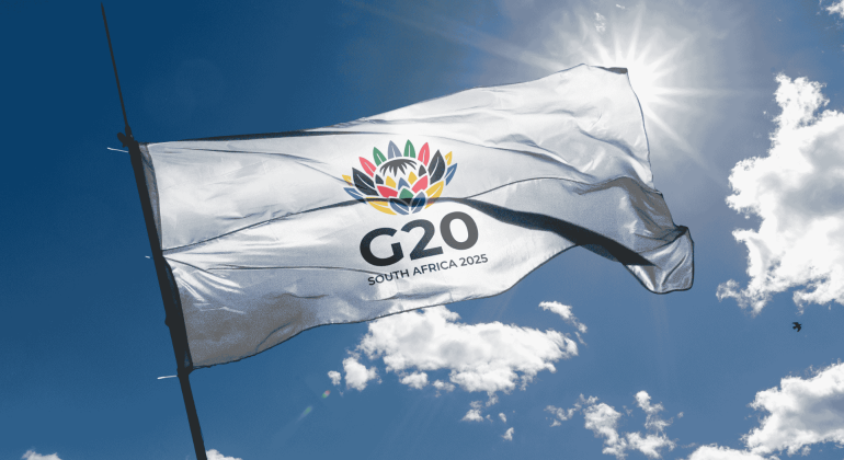 A white flag with a colorful flower emblem and the text G20 SOUTH AFRICA 2025 flutters against a sunny, blue sky, symbolizing new business opportunities on the global stage.