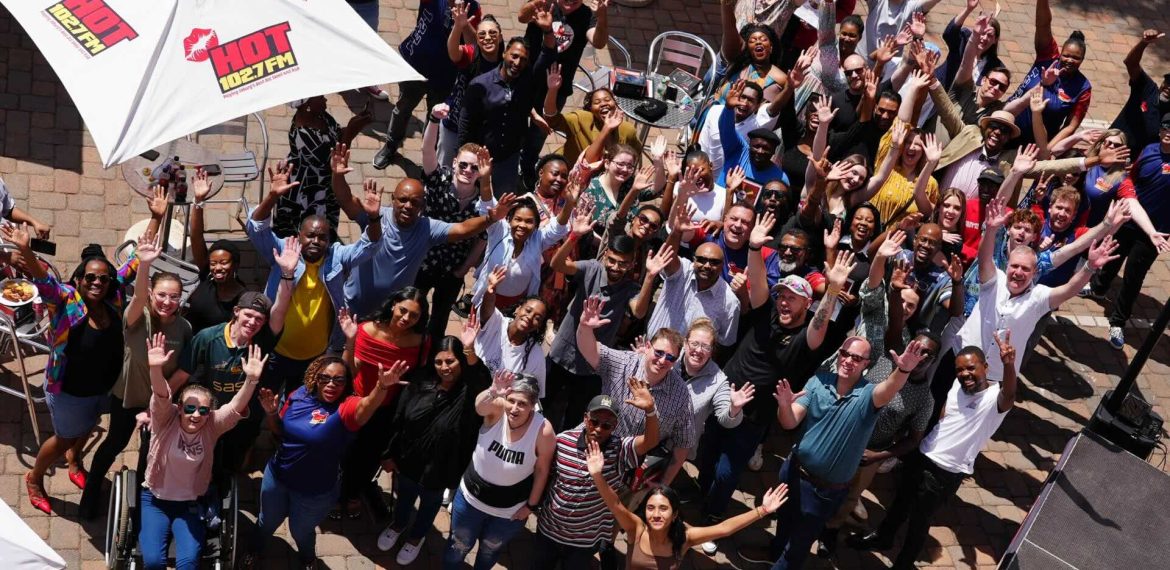A large, diverse group of people outdoors on a patio, smiling and waving up at the camera. Some are seated, most are standing under umbrellas with HOT 1027 FM logos. It’s a bright, sunny day filled with excitement and new business opportunities.