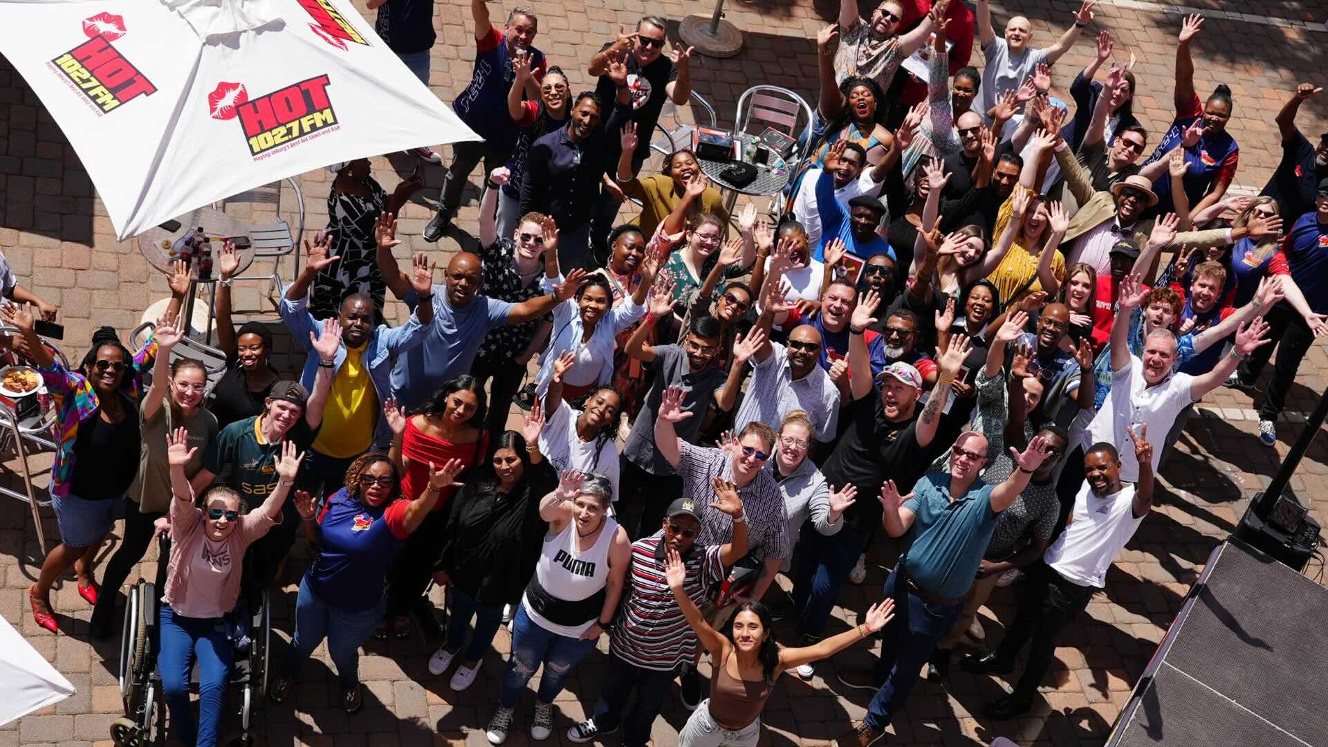 A large, diverse group of people outdoors on a patio, smiling and waving up at the camera. Some are seated, most are standing under umbrellas with HOT 1027 FM logos. It’s a bright, sunny day filled with excitement and new business opportunities.