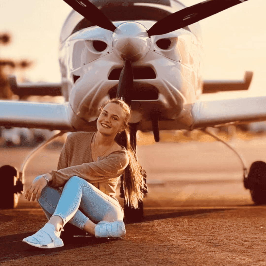 A young woman with long blonde hair sits smiling on the ground in front of a small white propeller airplane at sunset, reflecting on new business opportunities. She wears light blue jeans, a beige sweater, and white sneakers.
