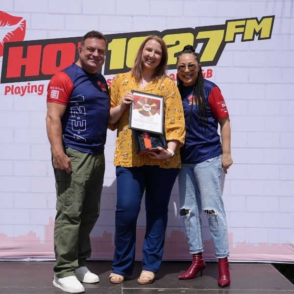 Three people stand smiling in front of a Hot 1027 FM banner. The woman in the center, holding a framed Radio Training Academy certificate, is flanked by two others as they pose together for a celebratory photo.