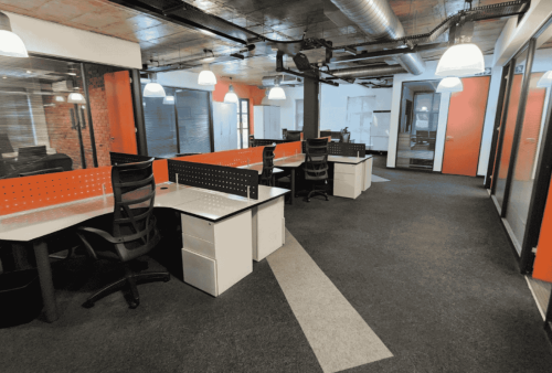 Modern open-plan office with several empty desks and black chairs, orange and white walls, glass partitions, carpeted floors, and exposed ceiling ducts—perfect for exploring new business opportunities.