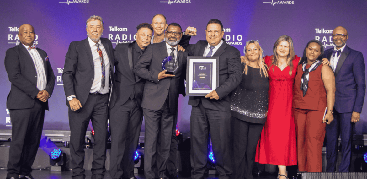 A group of eleven people in formal attire stand on stage at the Telkom Radio Awards near the Vaal Dam. One person holds a trophy while another holds a framed certificate, all smiling in front of a blue backdrop with the event logo.