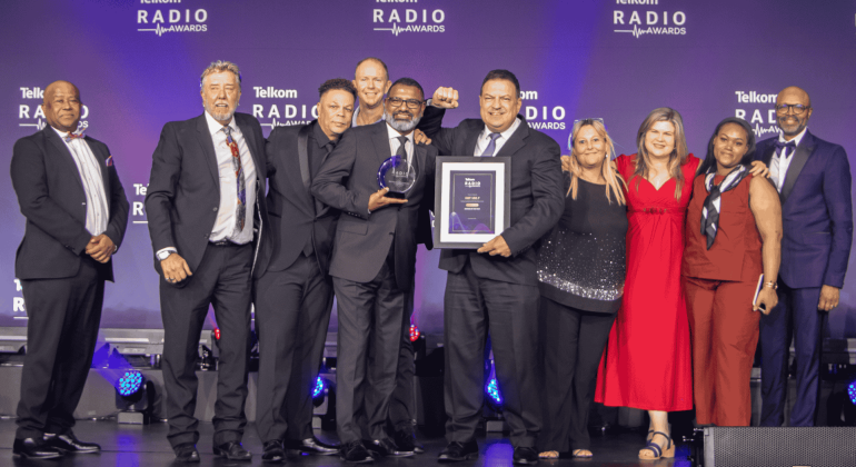 A group of eleven people in formal attire stand on stage at the Telkom Radio Awards near the Vaal Dam. One person holds a trophy while another holds a framed certificate, all smiling in front of a blue backdrop with the event logo.