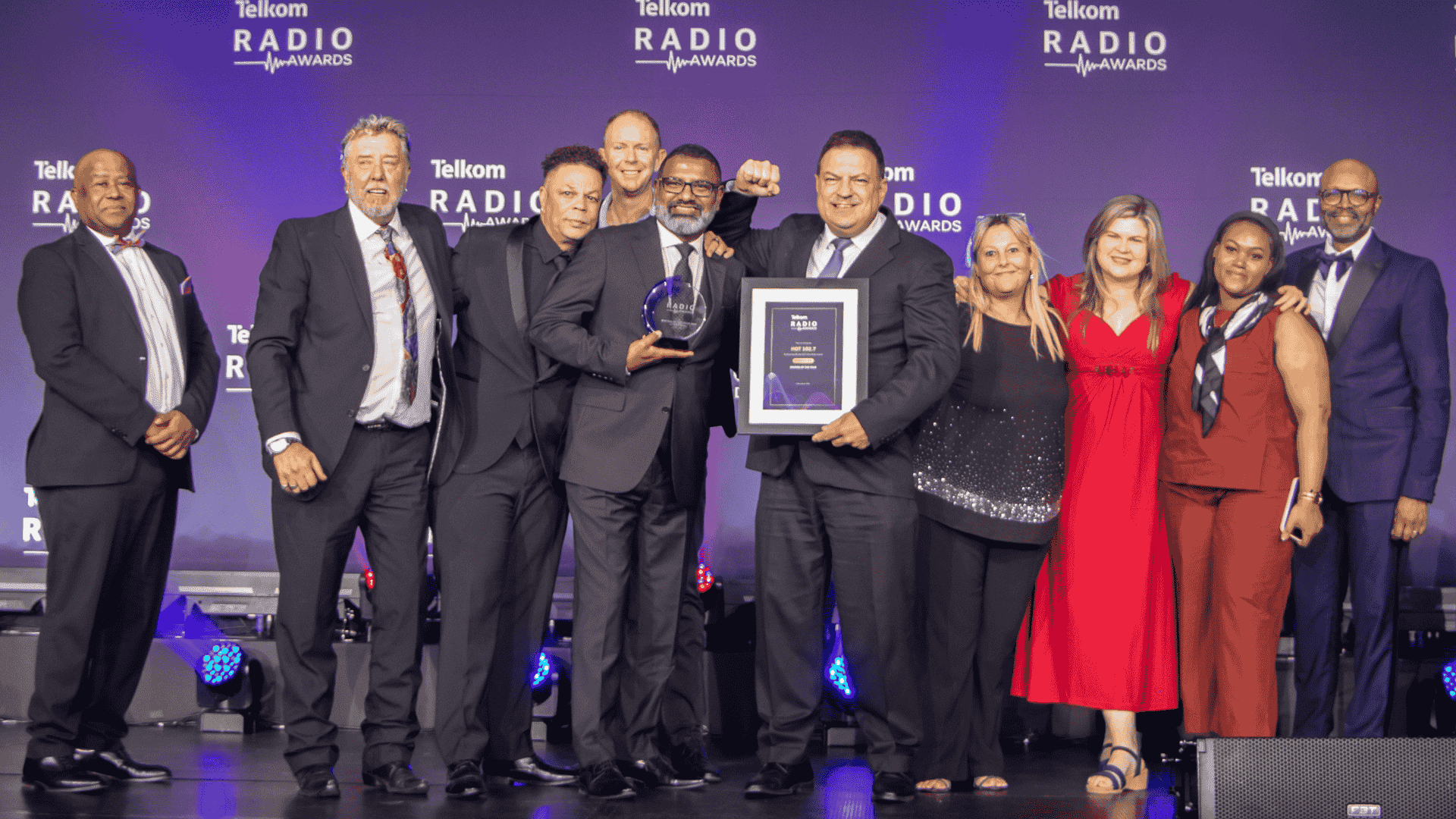 A group of eleven people in formal attire stand on stage at the Telkom Radio Awards near the Vaal Dam. One person holds a trophy while another holds a framed certificate, all smiling in front of a blue backdrop with the event logo.
