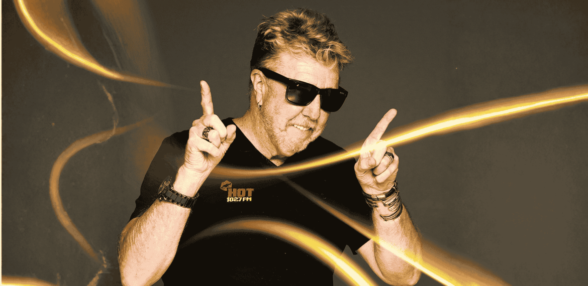 A man wearing sunglasses and a black HOT 102.7FM shirt smiles and points both index fingers upward, surrounded by swirling yellow light trails on a dark background, capturing the vibrant energy of Vaal Dam nightlife.