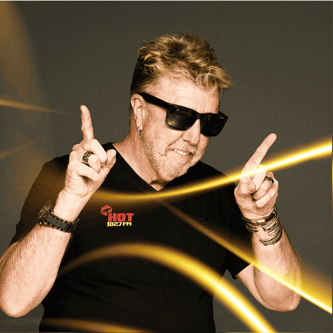 A man with light hair, wearing sunglasses and a black HOT 102.7 FM shirt, smiles and points both index fingers upward at the Vaal Dam. He is accessorized with rings and bracelets, with yellow light streaks in the foreground.