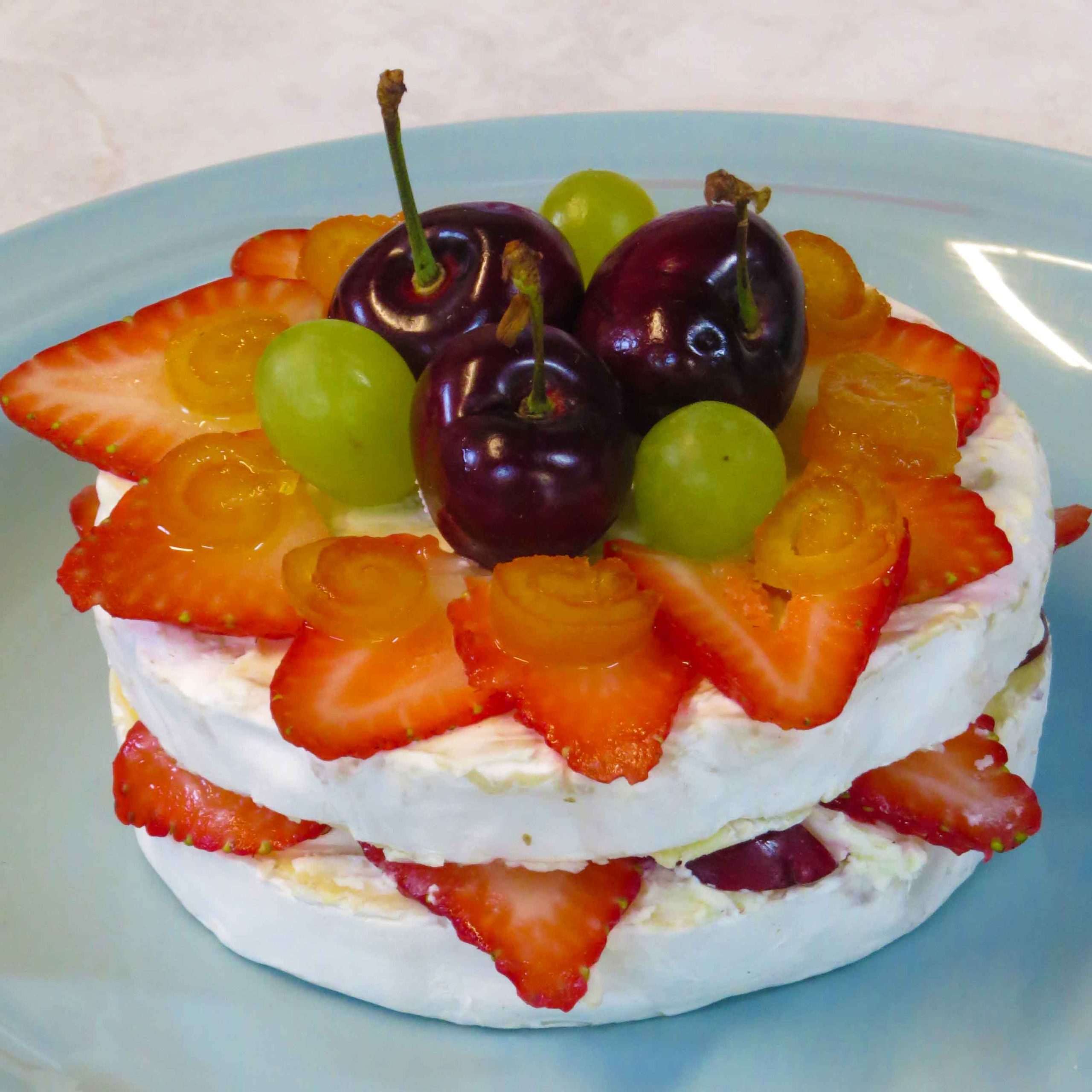 A round cheese wheel layered with sliced strawberries, garnished with cherries, green grapes, and small yellow fruit spirals, is beautifully displayed on a light blue plate—a true foodie’s delight.