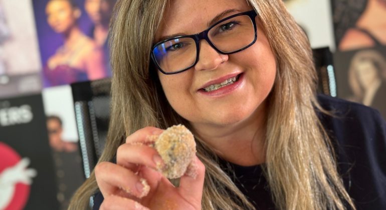 A woman with long blonde hair and glasses smiles at the camera, holding up a small breaded Matric snack. Blurred music posters are visible in the background.
