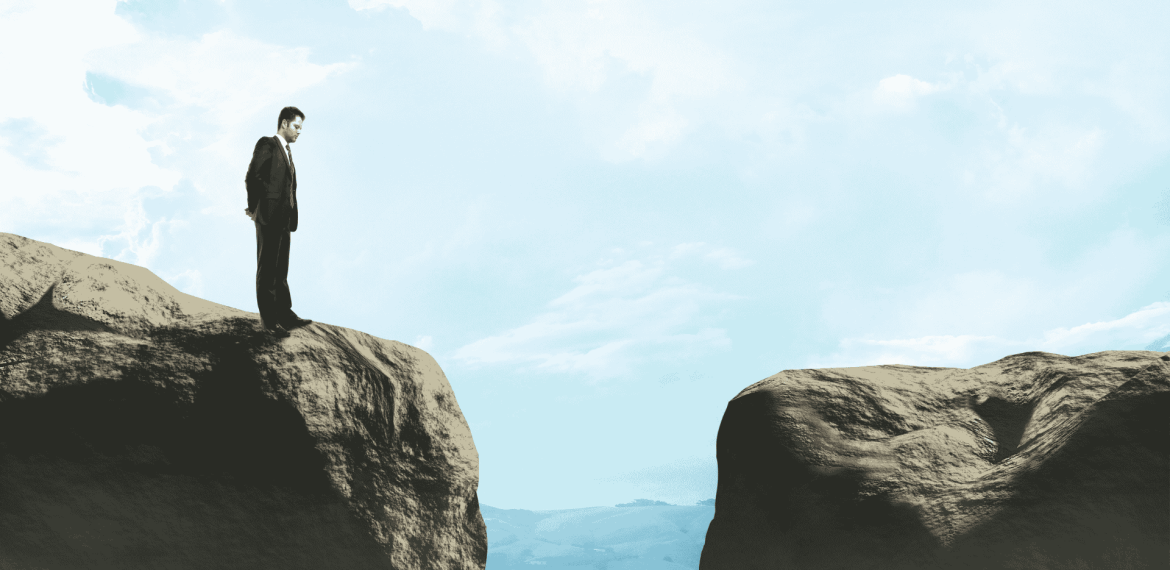 A man in a suit stands on the edge of a cliff, looking across a wide gap toward another rocky ledge under a blue sky with clouds, contemplating new business opportunities.