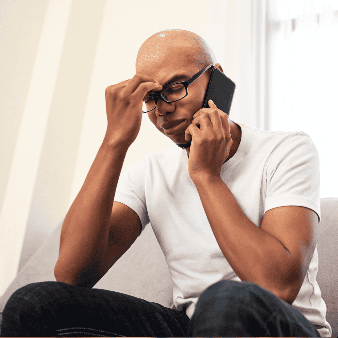 A man wearing glasses and a white t-shirt sits on a couch, holding a phone to his ear and pinching the bridge of his nose, appearing stressed or frustrated by business opportunities.
