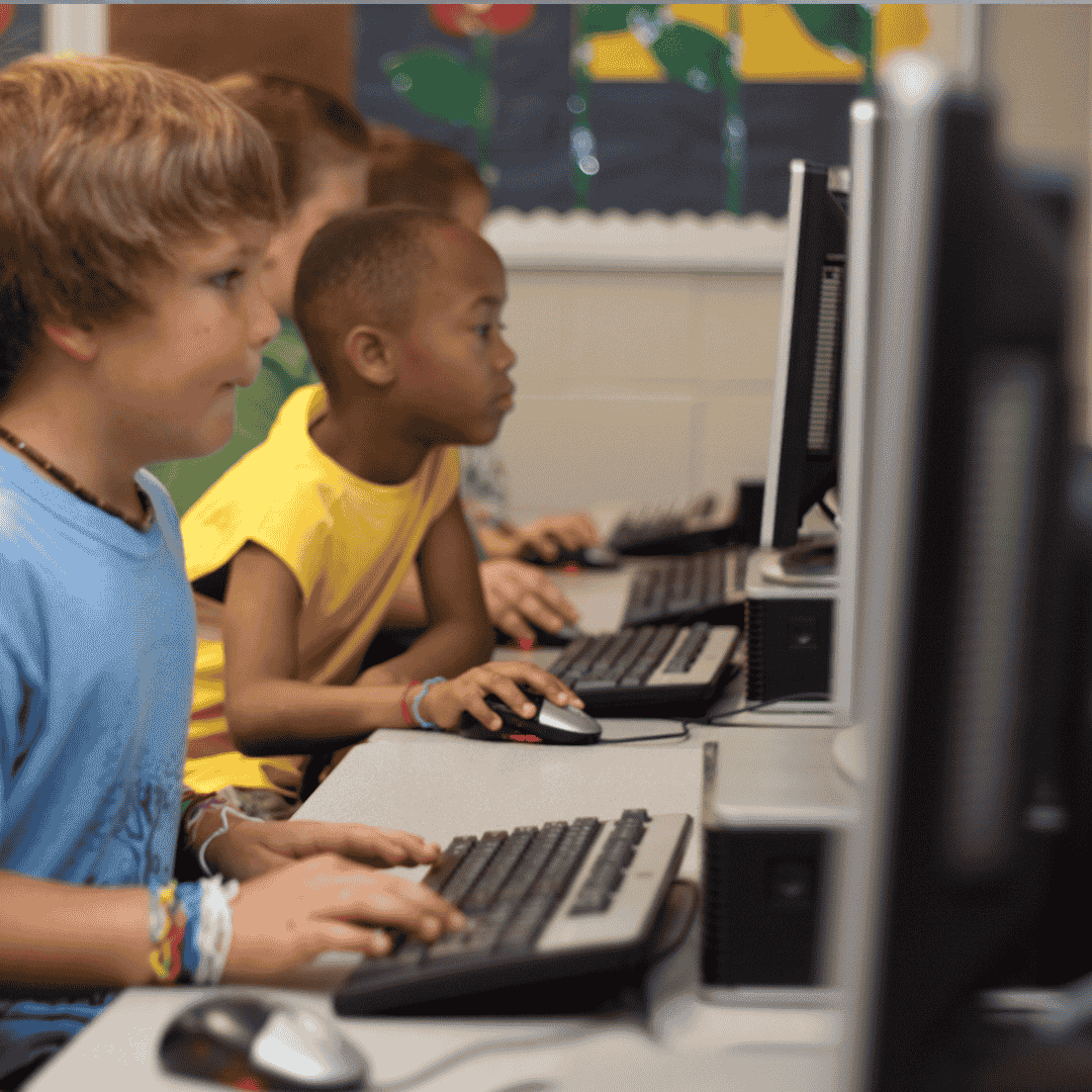 Children sit side by side at computers in a classroom, focused on their screens as they use keyboards and mice—developing valuable skills that will help them prepare for Matric. The background shows a bulletin board and classroom decorations.