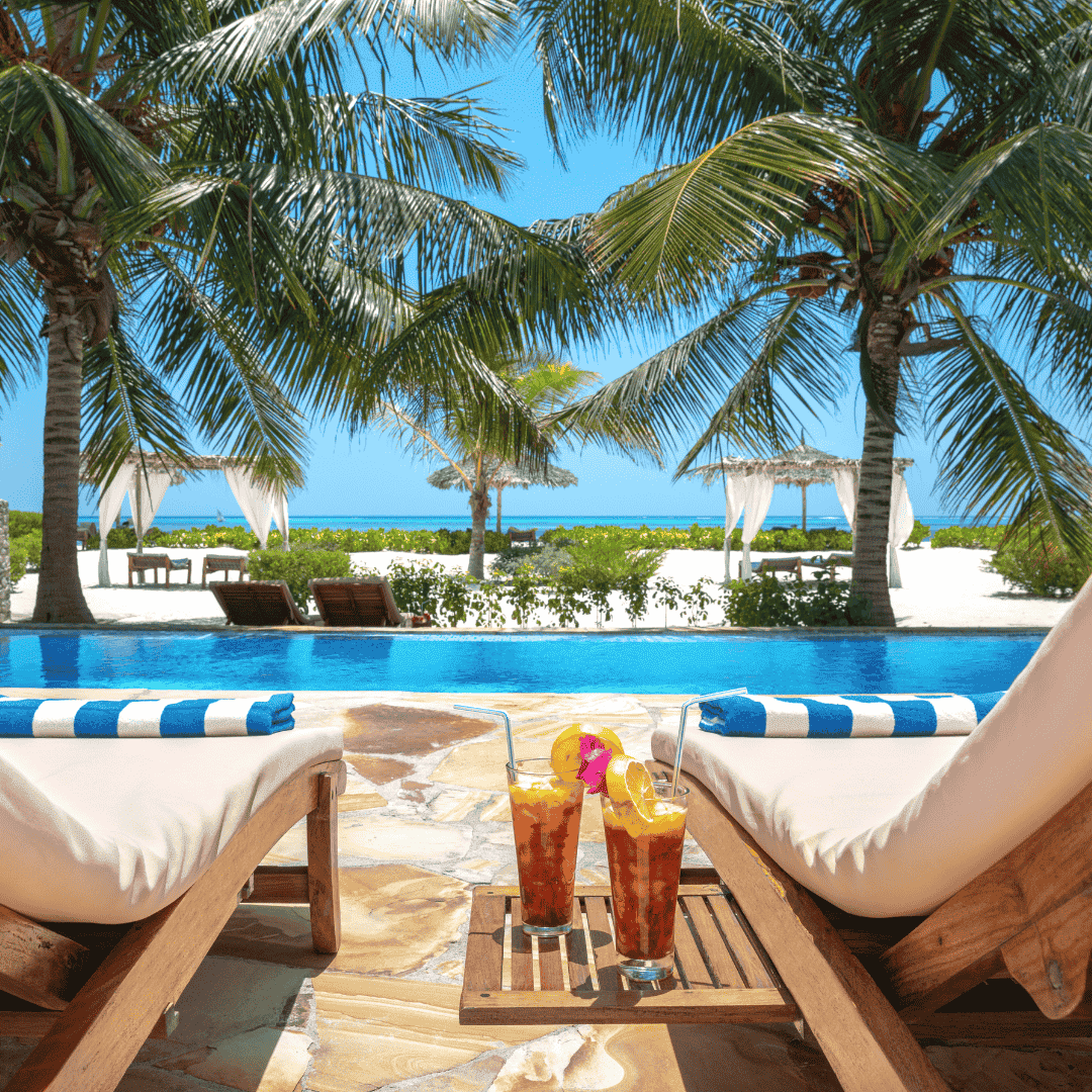 Two lounge chairs with striped towels face a pool, palm trees, and a sandy beach with cabanas. Two tropical drinks on a small wooden table evoke the perfect Matric summer vacation—a relaxing escape after exams.