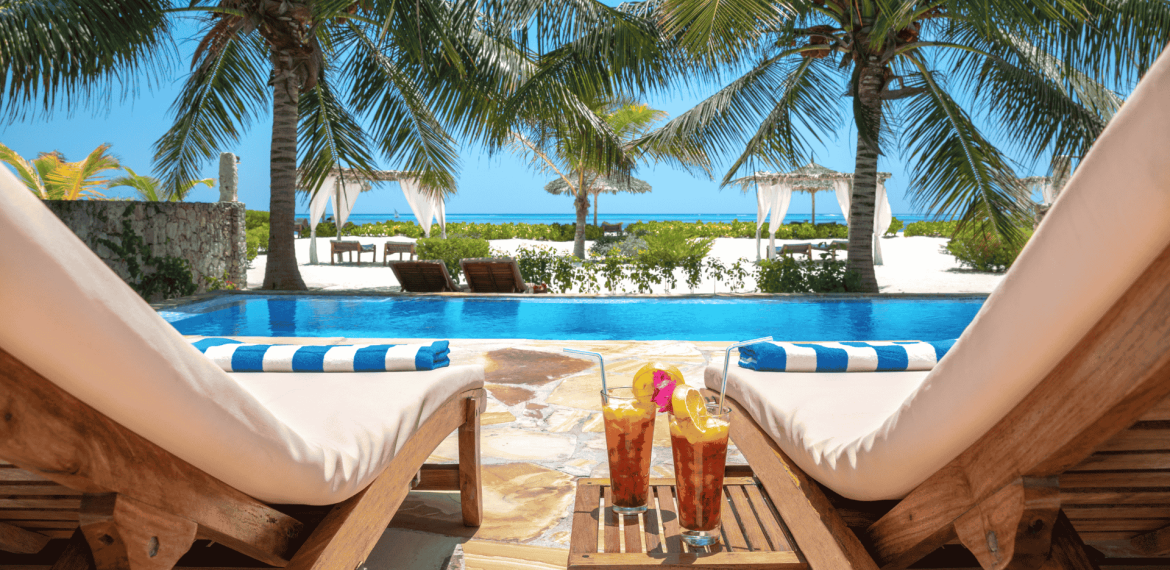 Two lounge chairs by a pool overlook a tropical beach with palm trees and turquoise ocean. Matric exams behind you, sip iced drinks with fruit garnish under sunny skies, relaxing by the white canopies and gentle waves.