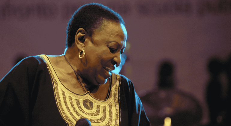 A woman wearing a black and gold traditional outfit smiles while holding a microphone on stage in Yeoville, with blurred figures and a purple-lit background behind her.