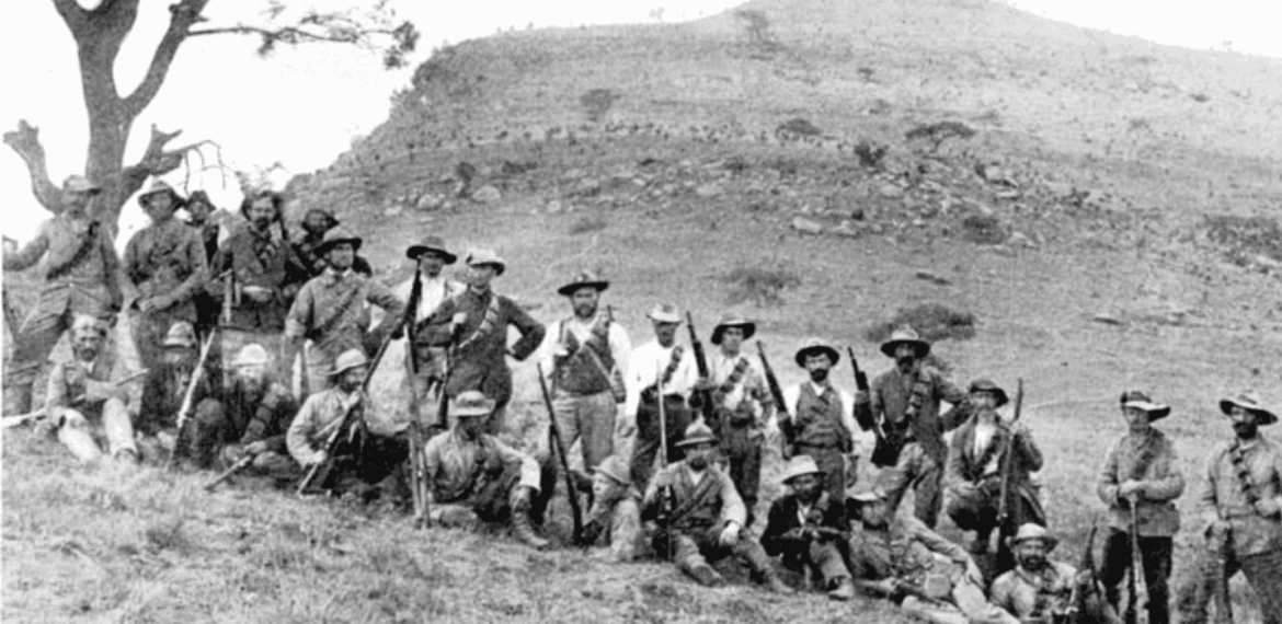 A group of men in hats and old-fashioned clothing pose with rifles on a grassy hillside near Yeoville; a lone tree stands to the left and a rugged hill rises in the background. The photo appears to be historical.