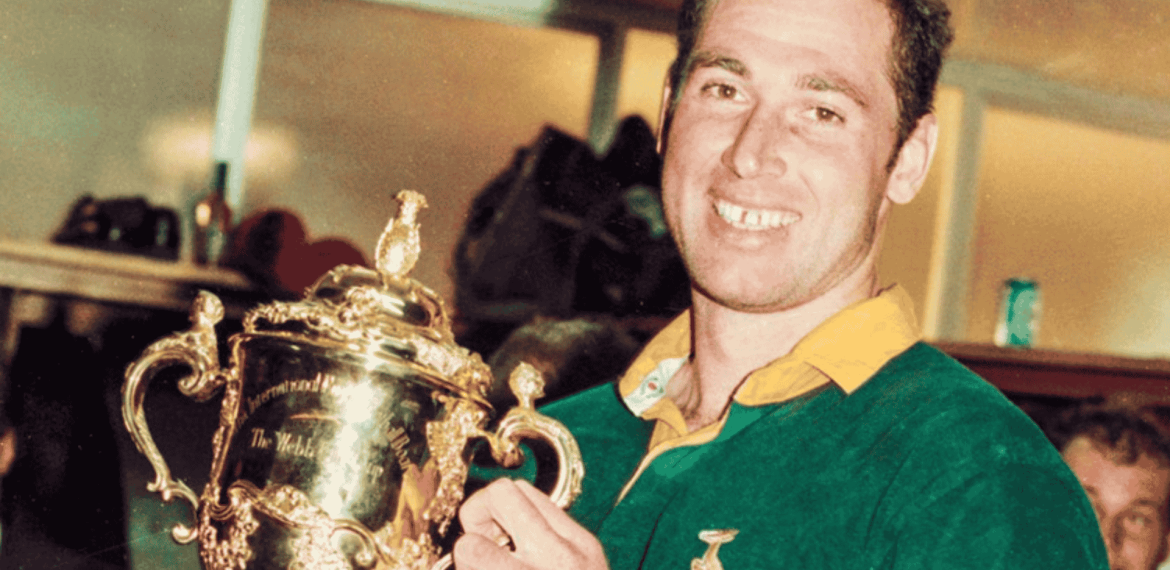 A smiling rugby player in a green jersey holds up a gold trophy cup, celebrating a cross-border victory indoors.