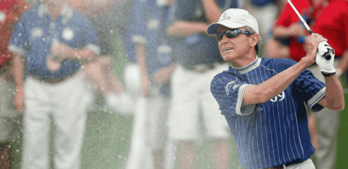 A golfer in a blue striped shirt and white cap swings a club, sending sand spraying into the air during a cross-border tournament, with several people in the blurred background watching.