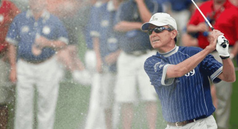 A golfer in a blue striped shirt and white cap swings a club, sending sand spraying into the air during a cross-border tournament, with several people in the blurred background watching.