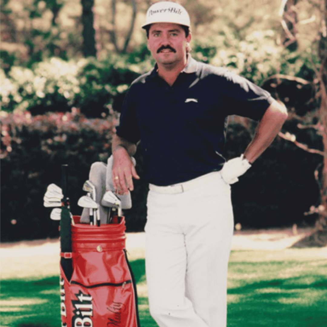 A man wearing a navy shirt, white pants, and a PowerBilt cap stands on a golf course, leaning on a red golf bag filled with clubs. The cross-border setting is highlighted by trees and greenery in the background.