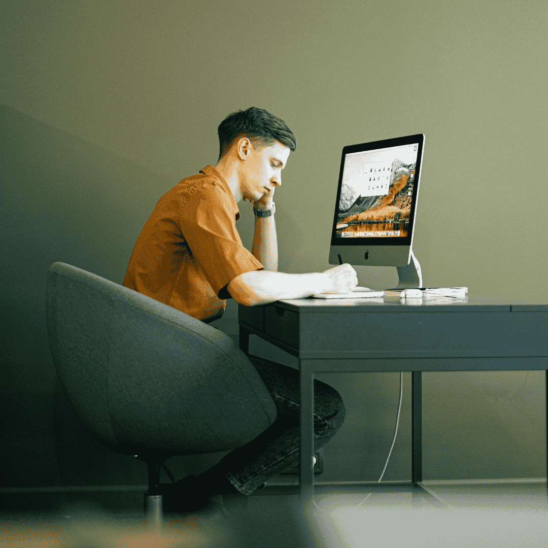 A man sits at a desk, exploring business opportunities as he writes on paper and looks at a computer monitor. He appears focused, with his head resting on one hand. The softly lit room contains papers, a keyboard, and the monitor.