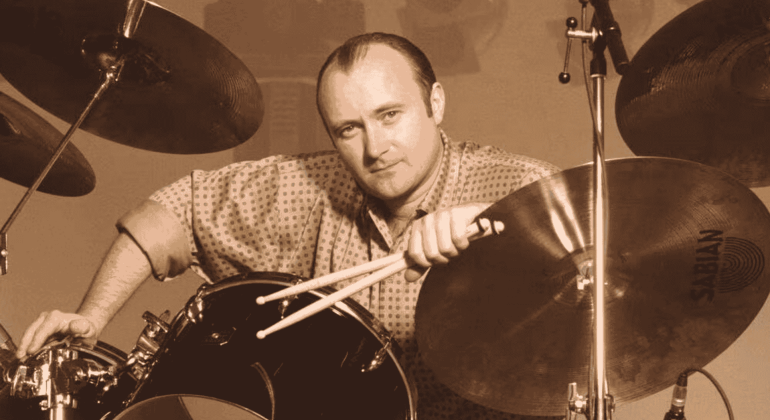 A man with a receding hairline and patterned shirt sits at a drum set, holding drumsticks and leaning on a Sabian cymbal, looking directly at the camera in a sepia tone—reminiscent of Phil Collins.
