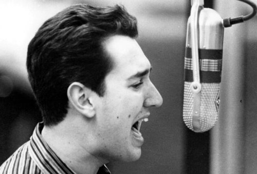 A young man with dark hair, wearing a striped shirt, sings passionately into a vintage microphone in this black and white photo, evoking the retro style of classic Blancmange performances.