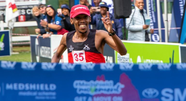 A male runner wearing a red cap and black-red outfit, bib number 35, smiles and raises his hand in celebration as he crosses the finish line at a race, just like he did last Christmas, with cheering spectators and banners in the background.