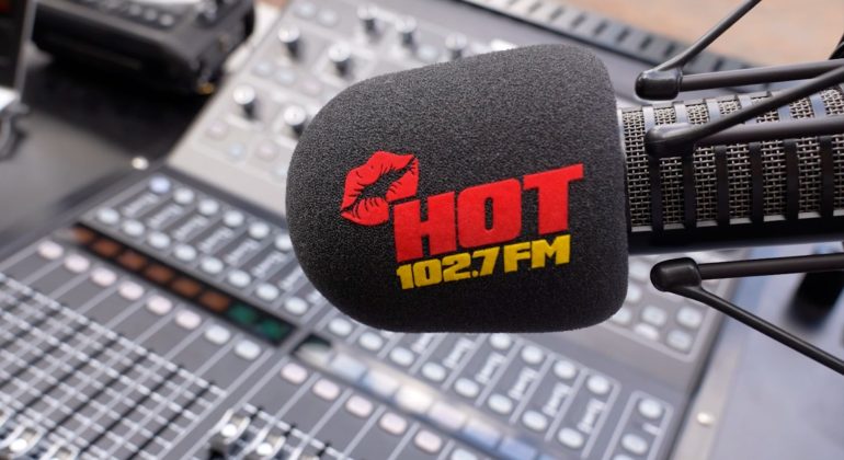 A microphone with a black foam cover displaying a red lipstick mark and the words HOT 102.7 FM in red and yellow, inspired by Miriam Makeba, positioned in front of a blurred audio mixing console.