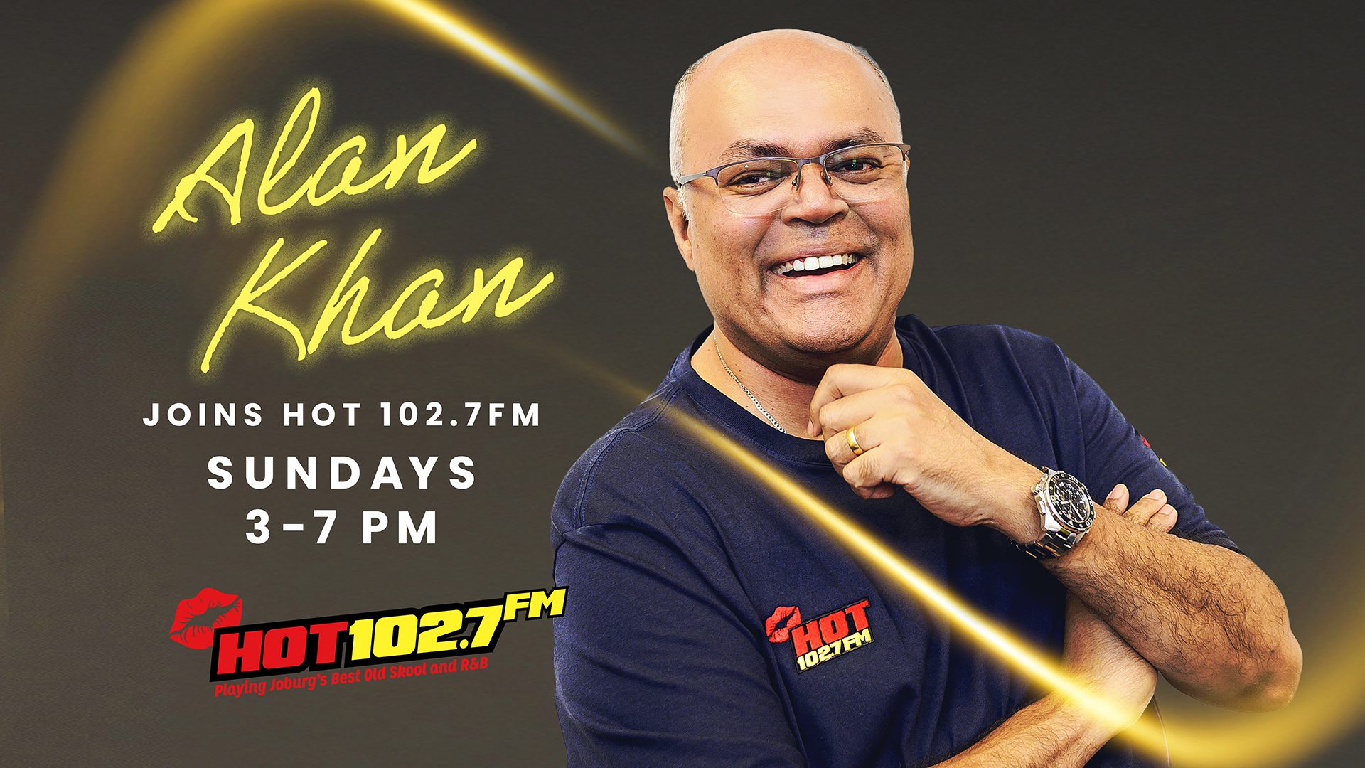 A smiling man in glasses and a black Hot 102.7 FM shirt stands with arms crossed. Text reads: Alan Khan joins HOT 102.7FM, Sundays 3–7 PM. The station logo is in the bottom left.