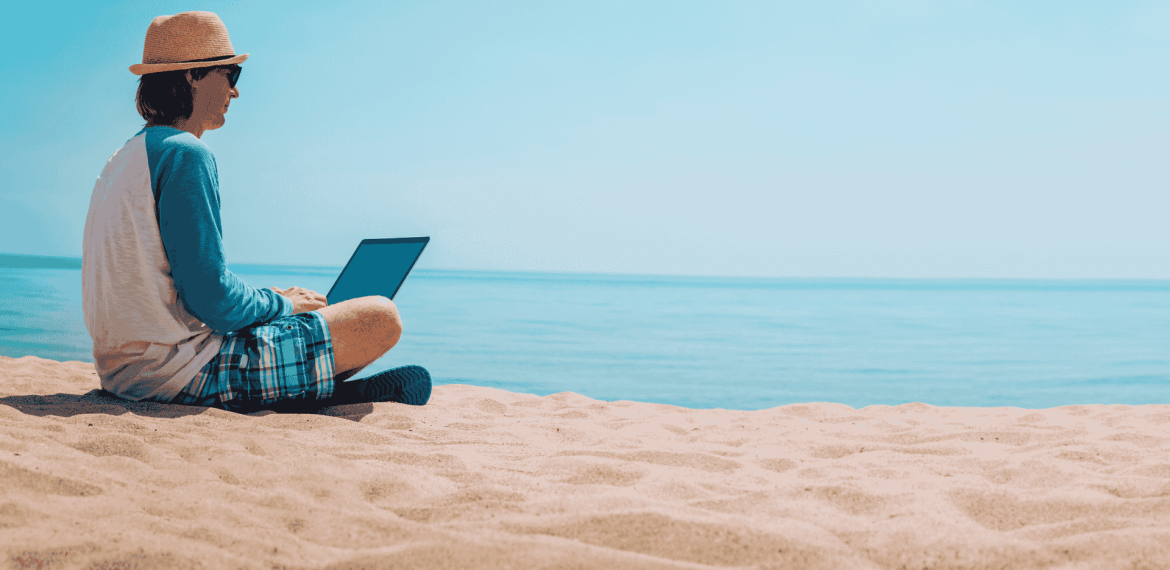 A person in a hat, sunglasses, and casual clothes sits on the sandy beach near the ocean, working on a Matric project on their laptop under a clear blue sky.