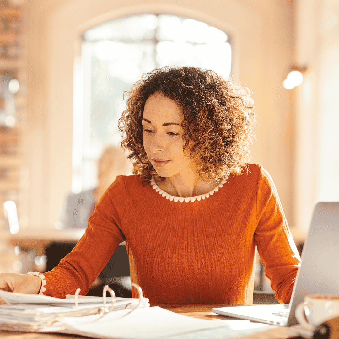 A woman with curly hair wearing an orange sweater sits at a desk, reviewing her Matric papers and working on a laptop in a bright, sunlit room.