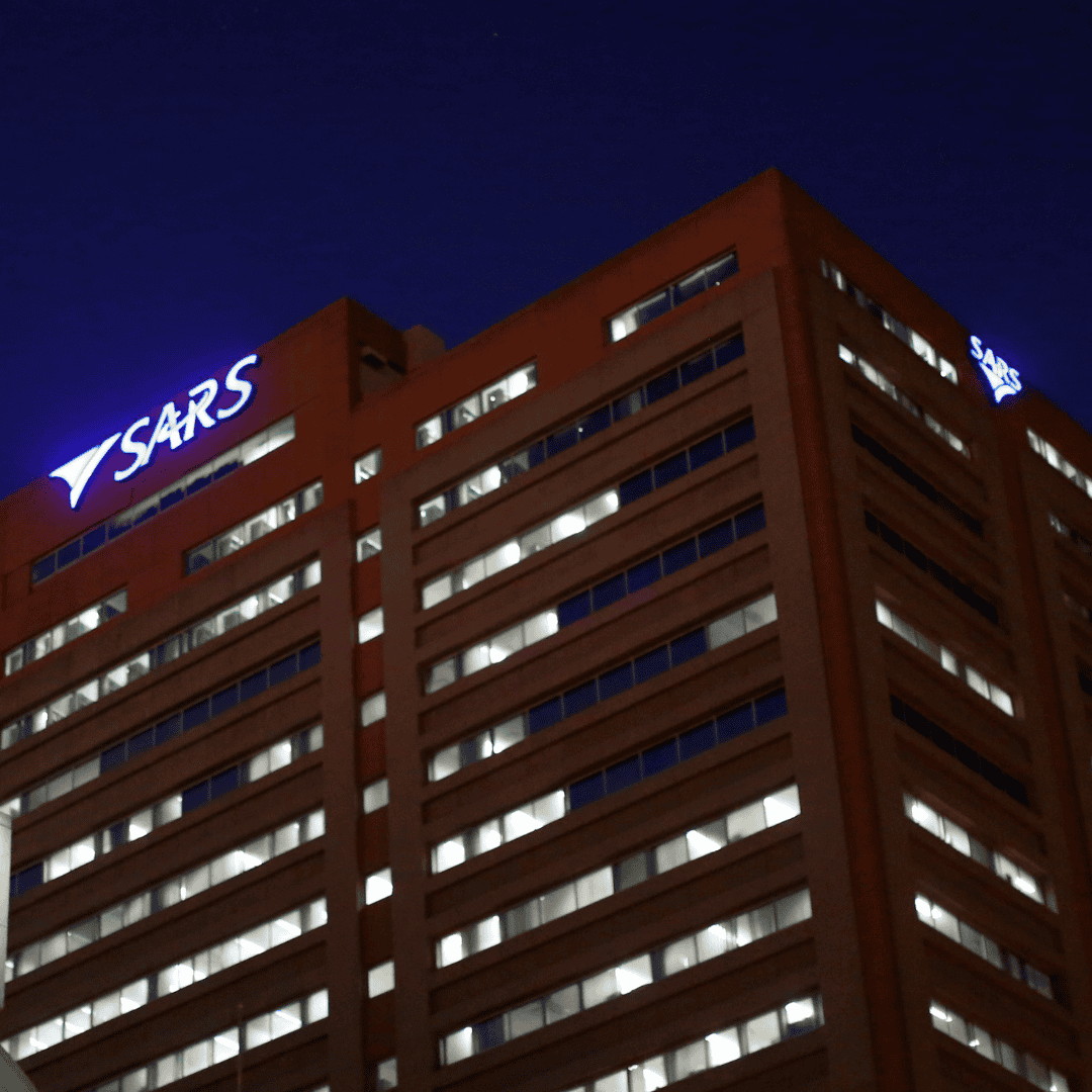 A multi-storey office building with many lit windows at night, featuring the illuminated SARS logo and name in white and blue on the top corner, stands tall like a beacon guiding Matric graduates towards new opportunities.