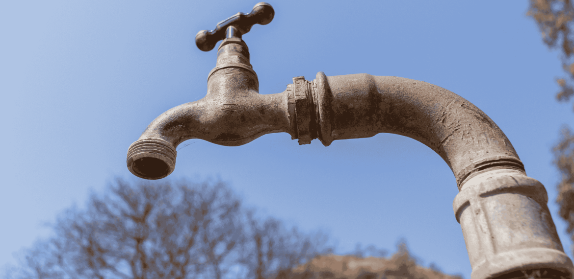 A close-up of an old, weathered water tap with no handle, set against a clear blue sky and blurred trees in the background, evokes the rustic aesthetic often featured in Die Kantoor.