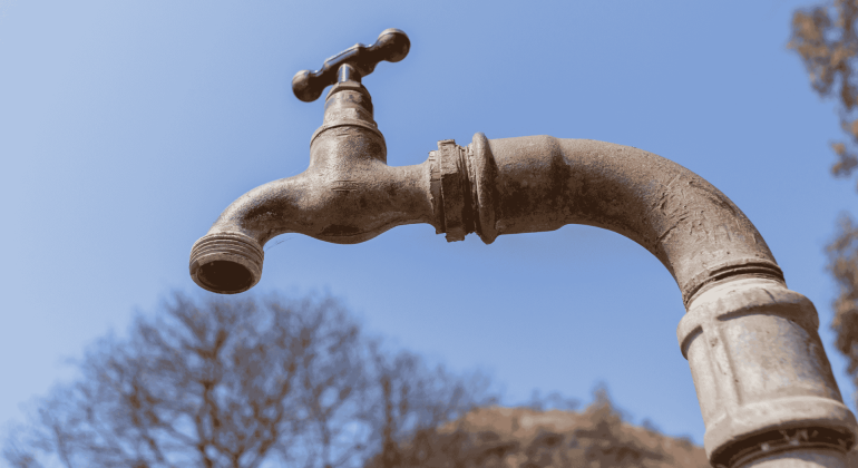 A close-up of an old, weathered water tap with no handle, set against a clear blue sky and blurred trees in the background, evokes the rustic aesthetic often featured in Die Kantoor.