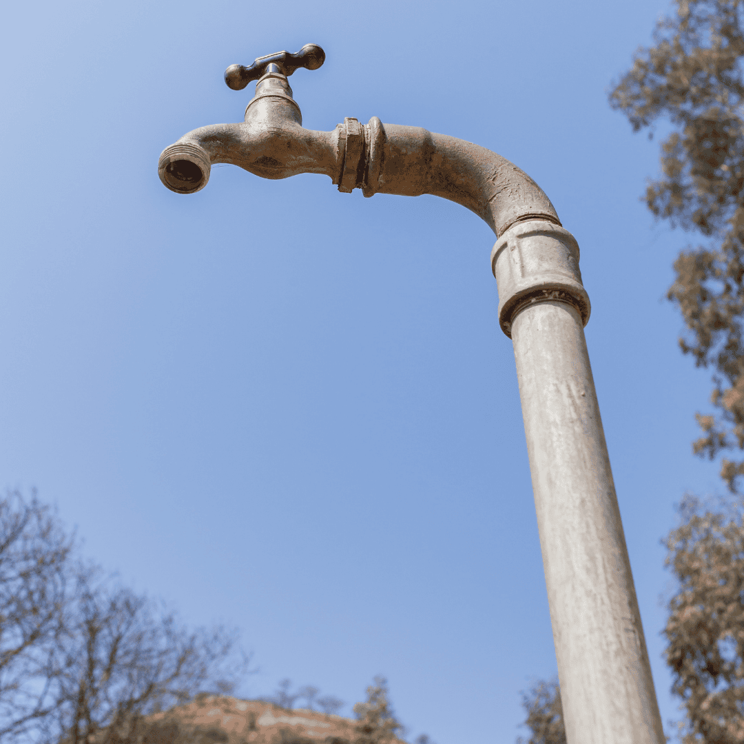 A weathered outdoor water tap on a tall metal pipe stands beneath the clear blue sky outside Die Kantoor, with blurry trees in the background.
