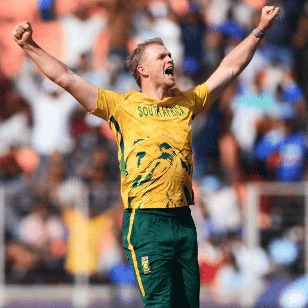 A South African cricket player in a yellow and green uniform celebrates with arms raised on the field, as spectators and blurred players look on—capturing the high-energy spirit of a Hot 1027 Breakfast moment.