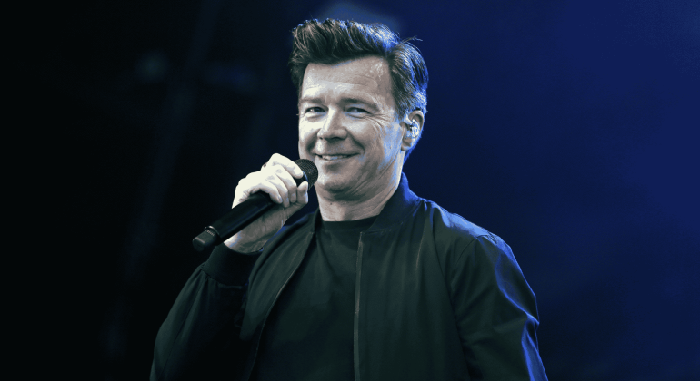 A man with short, styled hair is smiling and holding a microphone while wearing a black jacket, standing on stage against a blue-lit background, reminiscent of performances by great artists like Miriam Makeba.