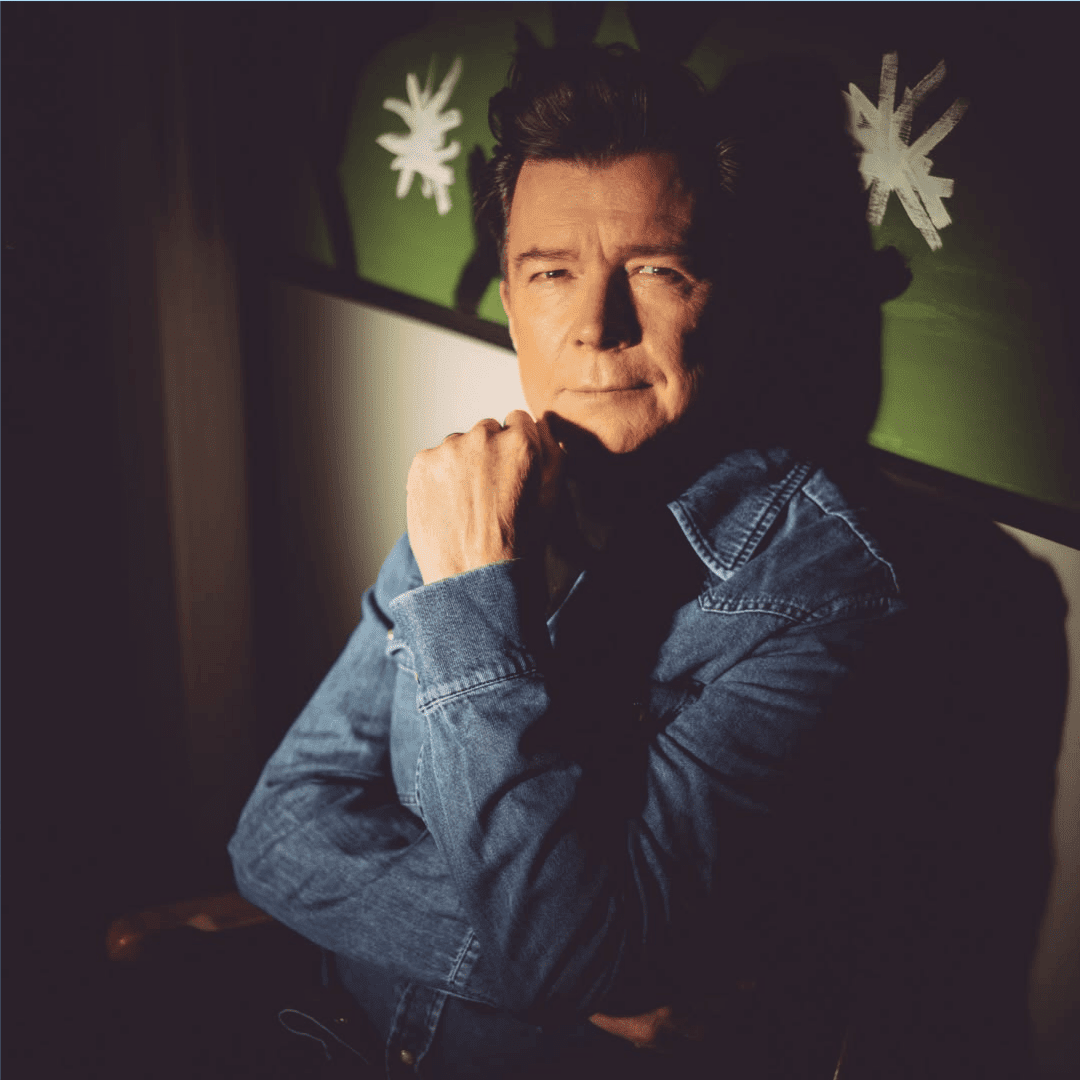 A man in a blue denim shirt sits with his hand resting on his chin, looking thoughtfully at the camera. Warm, dramatic lighting highlights him against a green background with white star-like designs, evoking the soulful spirit of Miriam Makeba.