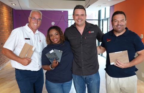Four people stand indoors, smiling at the camera. Three of them hold different flooring samples. The room has red brick and orange painted walls, with some construction wires and unfinished surfaces—a real hot 1027 breakfast vibe.