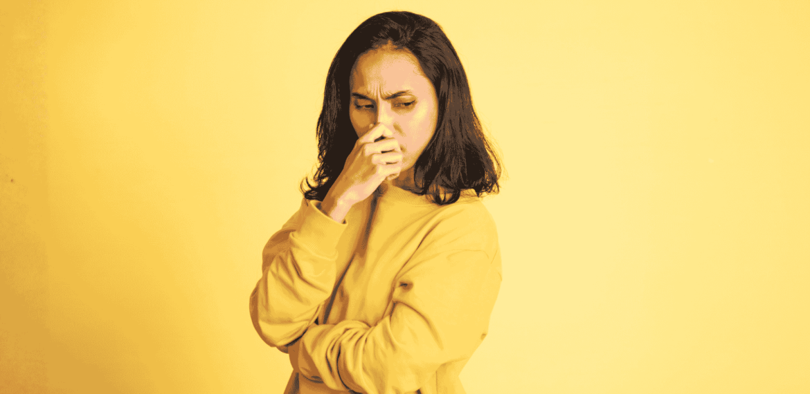 A woman in a yellow sweater stands against a yellow background, looking down with a worried or thoughtful expression—just like you might feel before coffee on the Hot 1027 Breakfast show—with her hand covering her mouth and her other arm crossed.