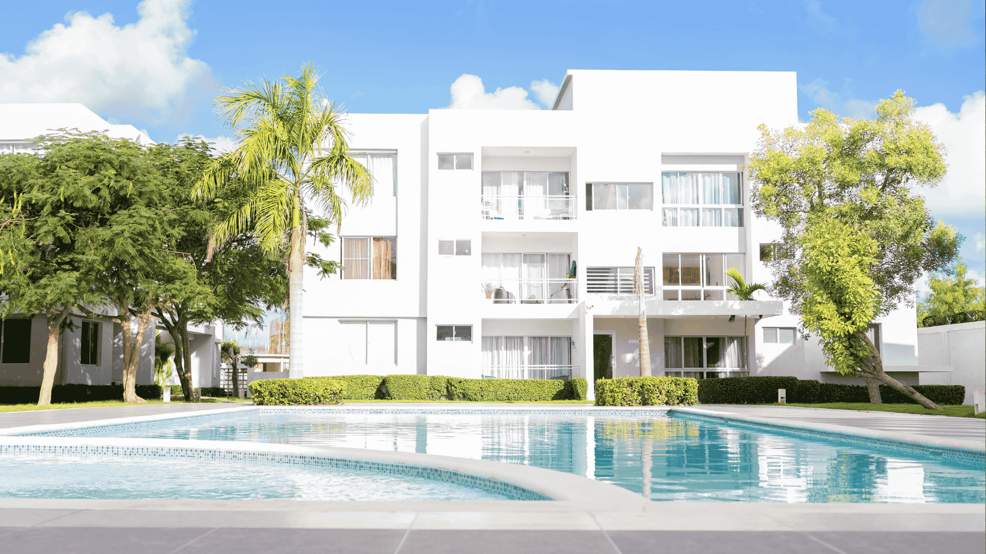 Modern white apartment building with large windows and balconies, surrounded by green trees and bushes, with a clear blue swimming pool in the foreground—perfect for enjoying your hot 1027 breakfast under a bright, sunny sky.