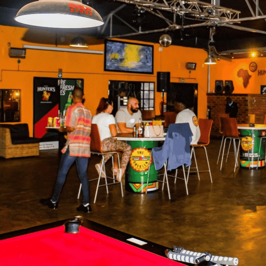 A lively bar with people sitting at barrel-style tables, a red pool table in the foreground, and yellow-orange walls decorated with posters, TVs, and a hot 1027 breakfast promo. The social atmosphere feels upbeat and relaxed as someone walks by.