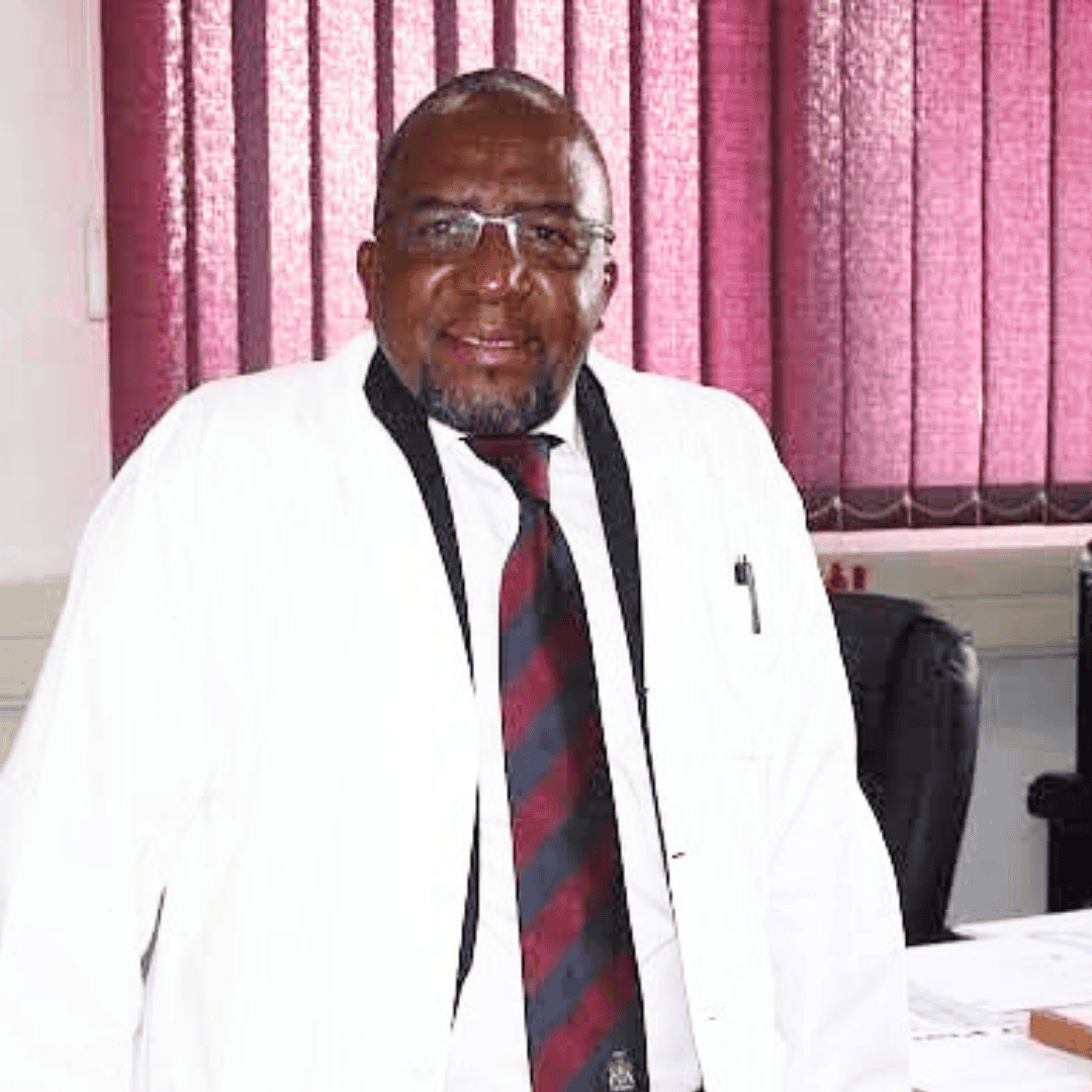 A man wearing glasses, a white coat, and a striped tie stands in an office with purple blinds and a desk in the background, ready to discuss the latest news from the Hot 1027 Breakfast show.