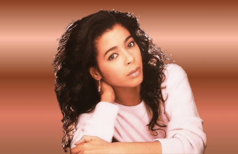 A woman with long, curly dark hair and a light pink sweater rests her head on one hand, looking calmly at the camera against a soft brown gradient background—perfectly capturing the relaxed vibe of a Hot 1027 Breakfast morning.